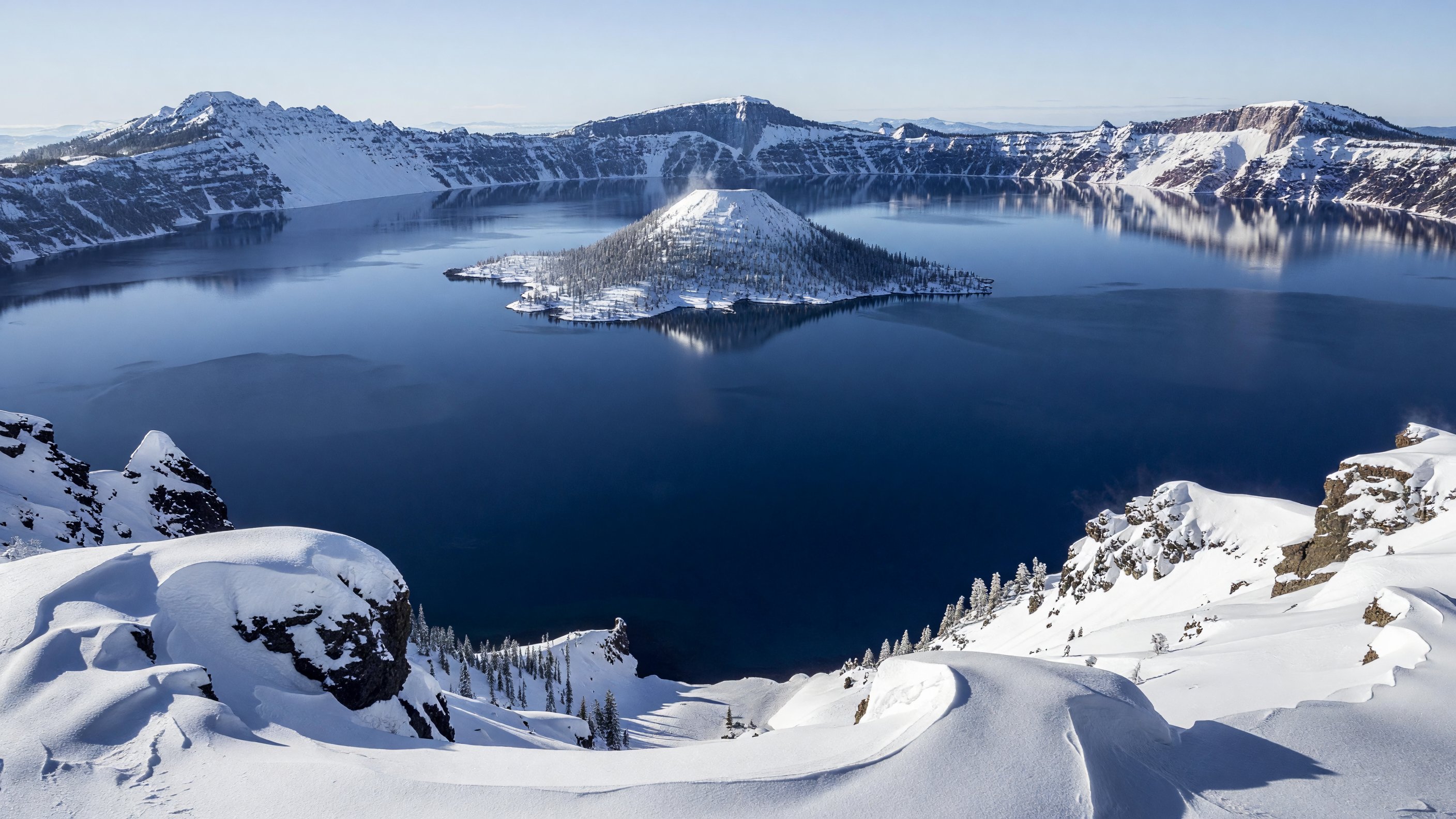 Crater Lake in winter — snow-blanketed caldera rim with the deep blue lake and Wizard Island visible below