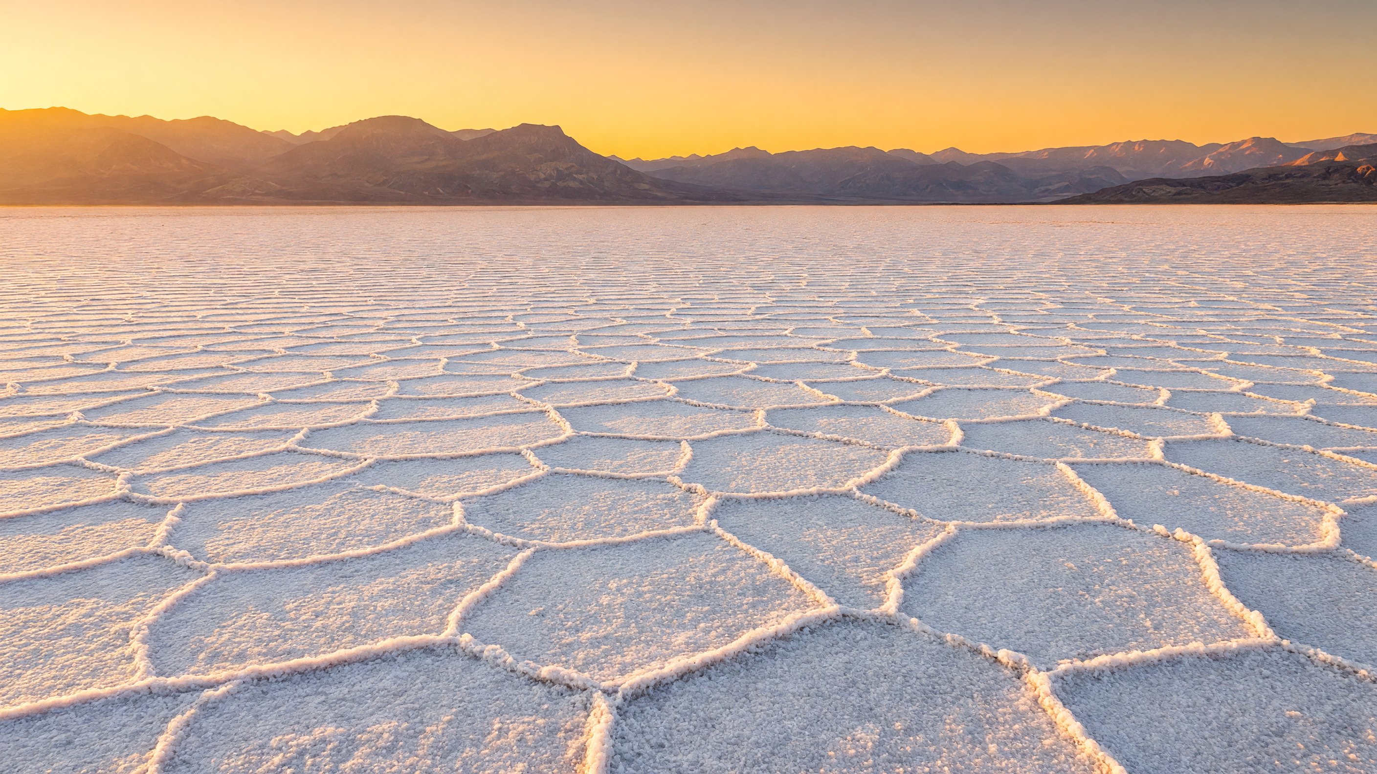 Wide panoramic view of Badwater Basin salt flats at Death Valley National Park at golden hour, crystalline white salt polygons extending to distant mountain ranges under a warm amber sky