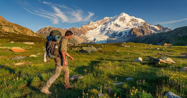 A backpacker with a loaded pack crossing a high alpine meadow with a glacier-capped peak in the background, late afternoon sun