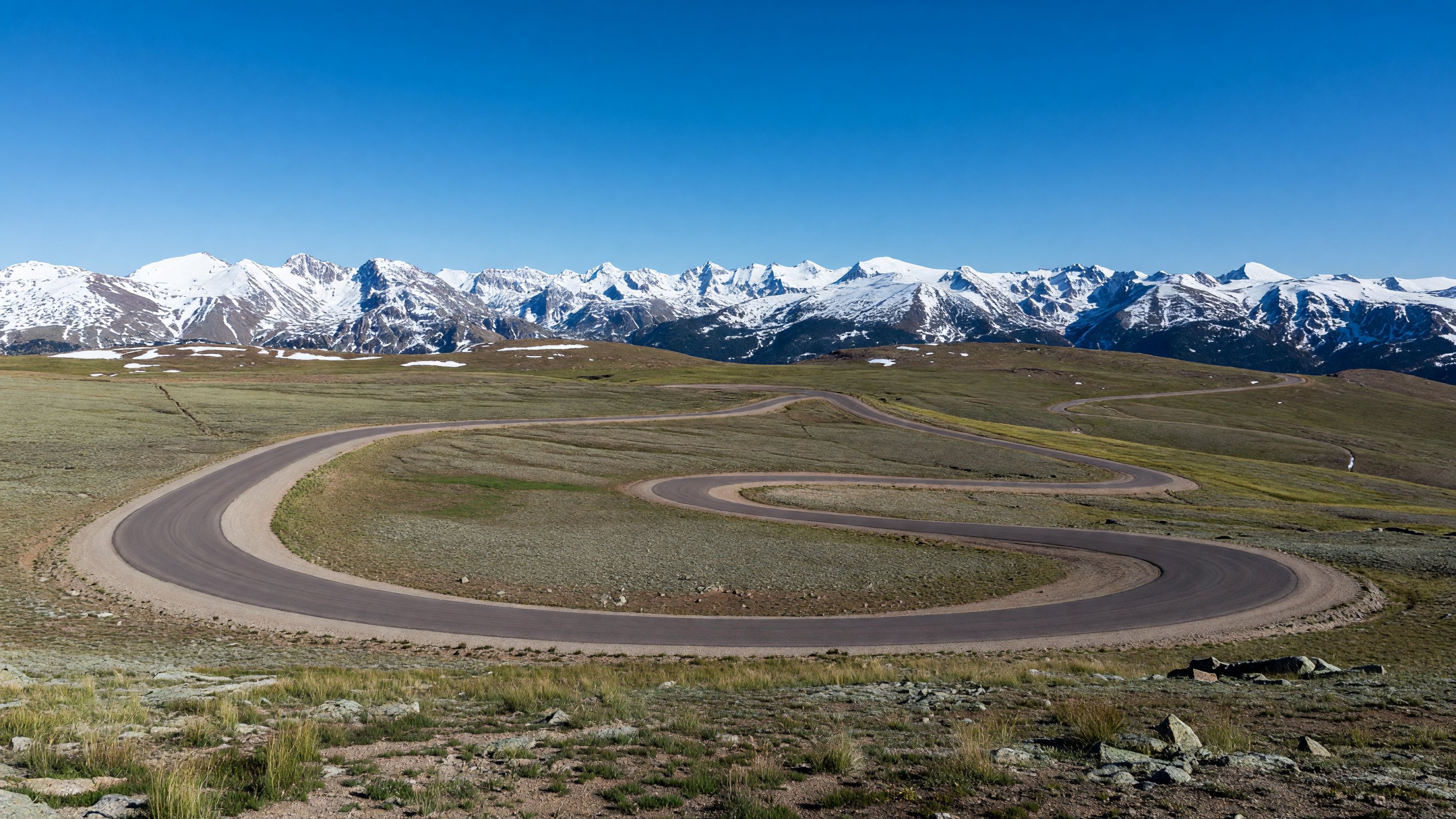 Trail Ridge Road winding through alpine tundra above treeline at Rocky Mountain National Park, Colorado, with a panorama of snow-capped peaks under a deep blue sky