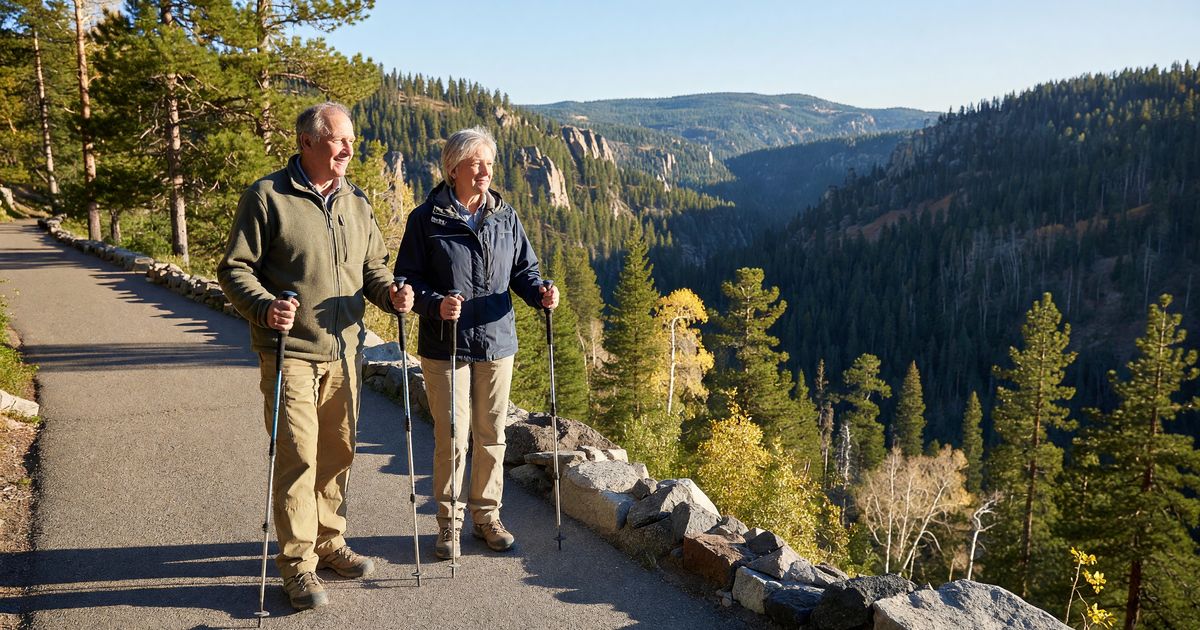 Two senior travelers with trekking poles on a paved overlook trail at a national park, late morning light, forested canyon below