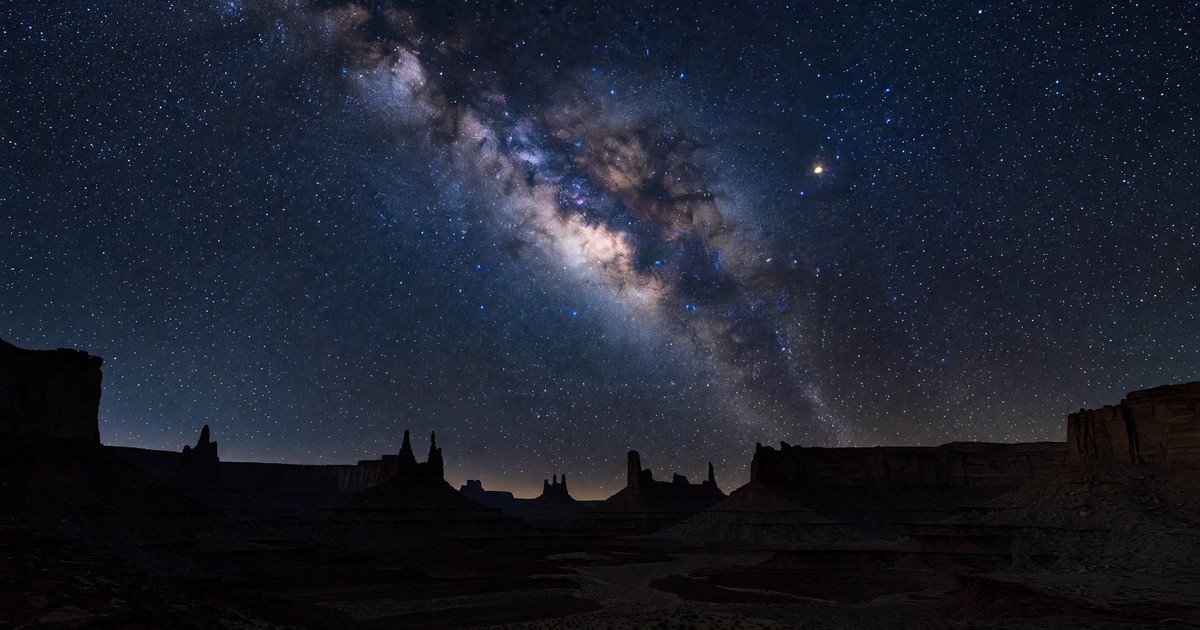 Wide-angle night sky photograph of the Milky Way galactic core arching over a silhouetted desert canyon landscape, with thousands of stars visible against a deep blue-black sky