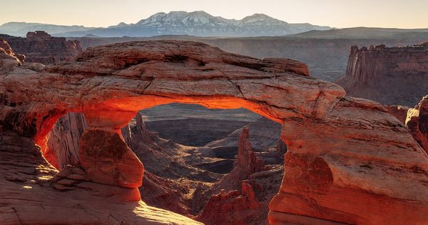 Mesa Arch at Canyonlands at sunrise — the underside of the sandstone arch glowing orange-red as first light fires through from the canyon below, with the La Sal Mountains visible in the background