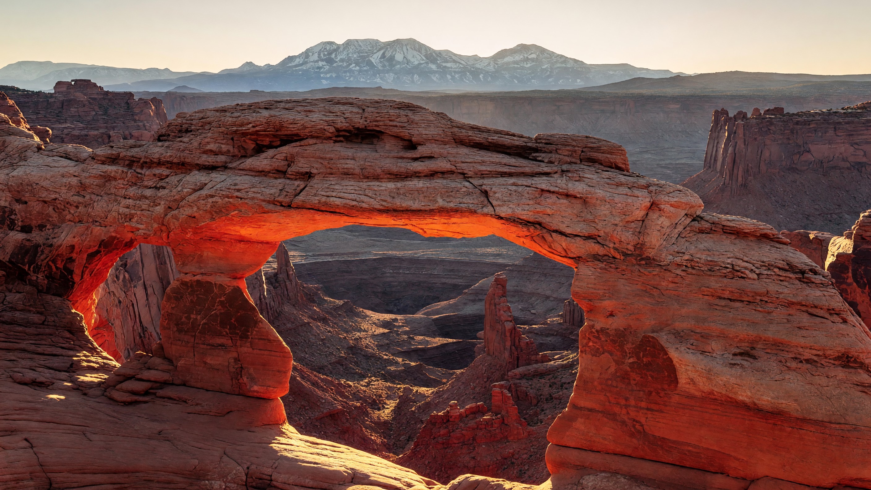 Mesa Arch at Canyonlands at sunrise — the underside of the sandstone arch glowing orange-red as first light fires through from the canyon below, with the La Sal Mountains visible in the background