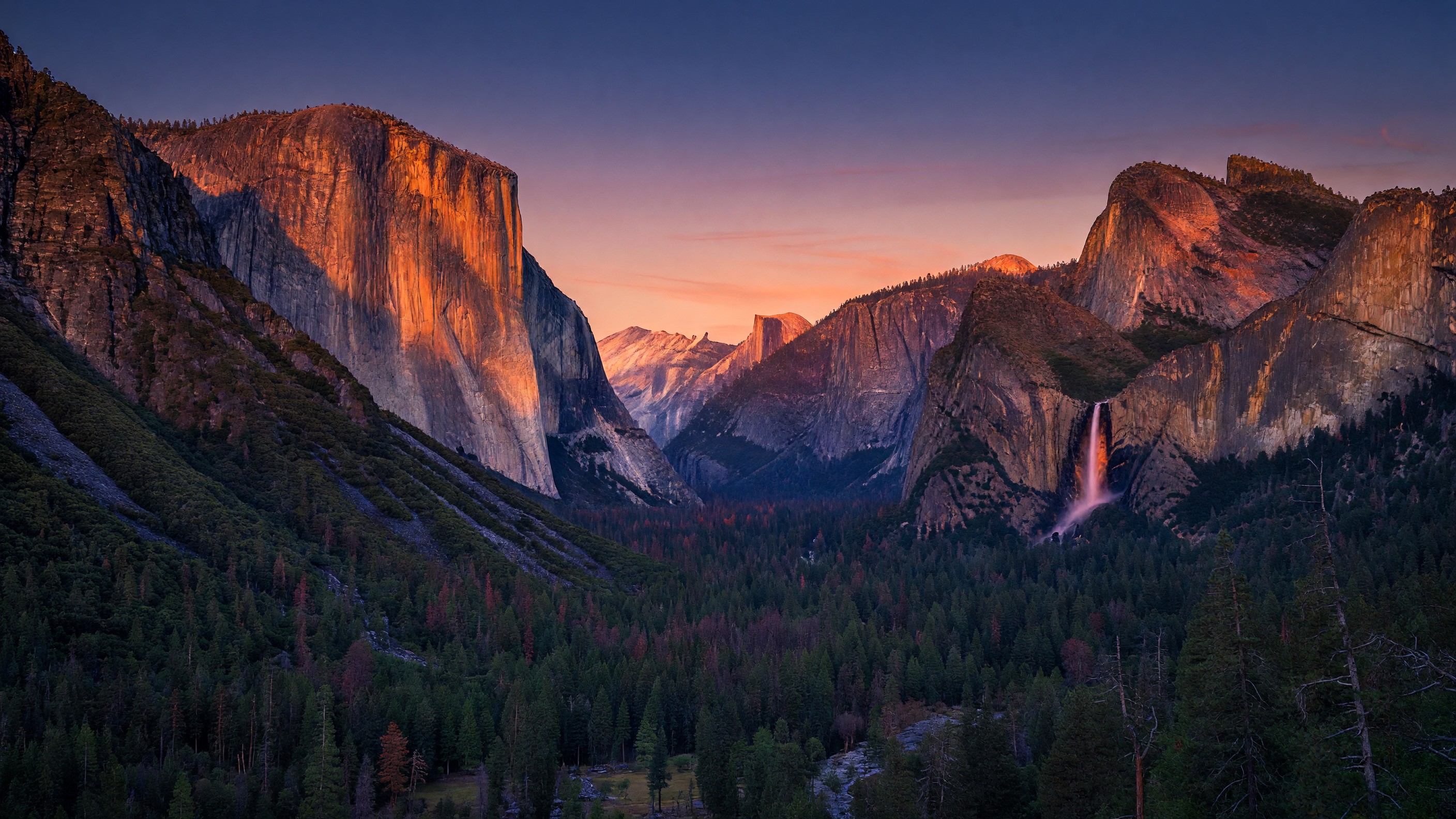 Tunnel View at Yosemite at sunset — El Capitan and Half Dome bathed in deep orange alpenglow, Bridalveil Fall catching the last warm light, Yosemite Valley floor in deep blue shadow below